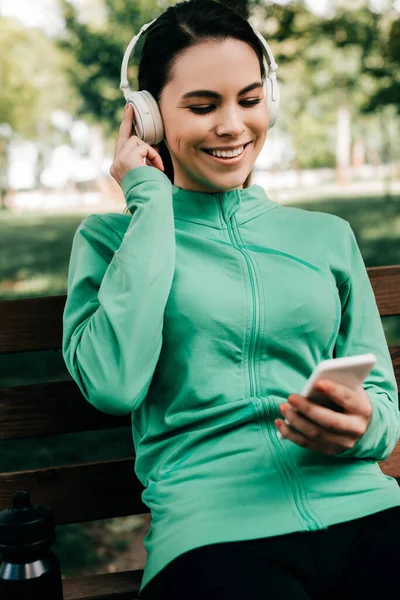 Selective Focus Smiling Sportswoman Listening Music Headphones Using Smartphone Bench — Stock Photo, Image