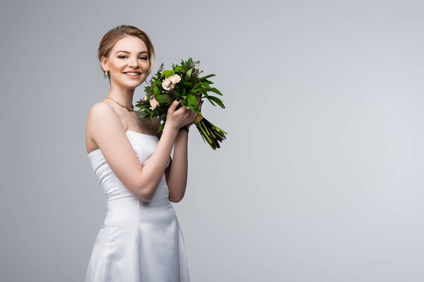 positive bride in white dress holding wedding flowers isolated on grey 