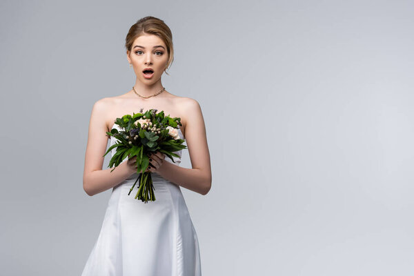 shocked bride in white wedding dress holding flowers isolated on grey 