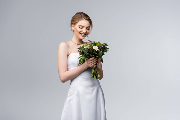young and happy bride in white dress holding bouquet of flowers isolated on grey 