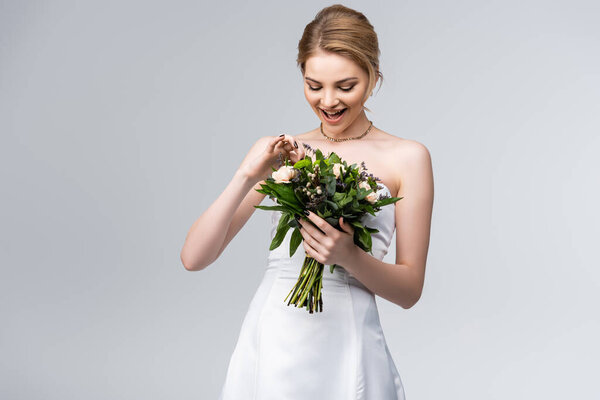 young and excited bride in white dress touching bouquet of flowers isolated on grey 