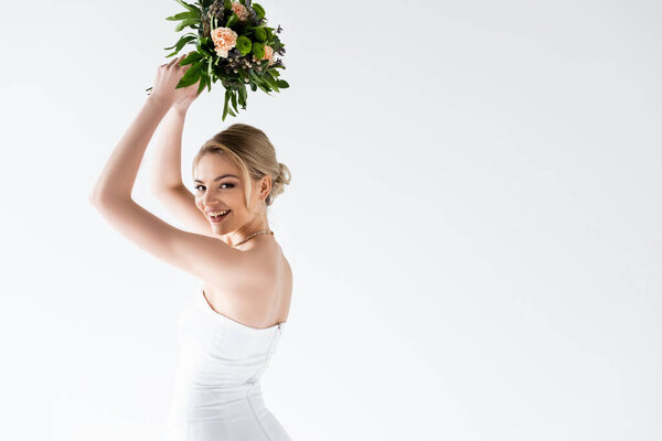 young and cheerful bride in elegant wedding dress holding flowers above head isolated on white 