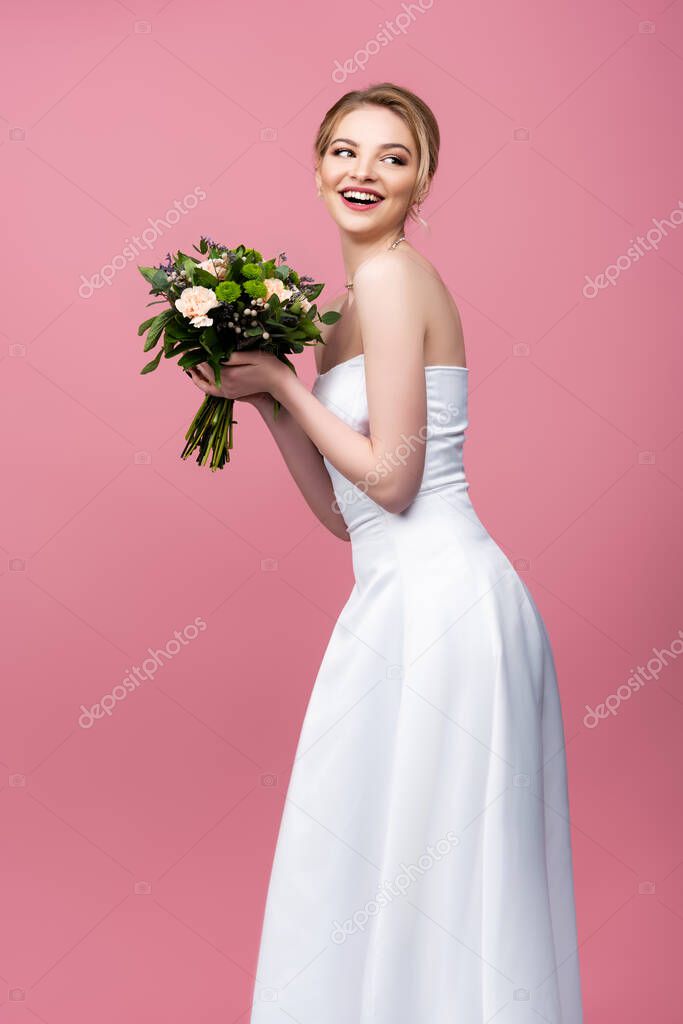 cheerful bride in white wedding dress holding flowers and looking away isolated on pink 