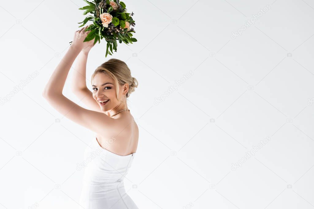 young and cheerful bride in elegant wedding dress holding flowers above head isolated on white 