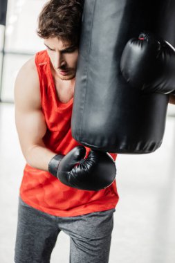 Spor giyim ve boks eldivenleri giymiş yorgun bir adam spor salonunda kum torbasına dokunuyor. 