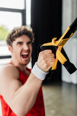 selective focus of sportive man screaming while working out with elastics in gym  clipart