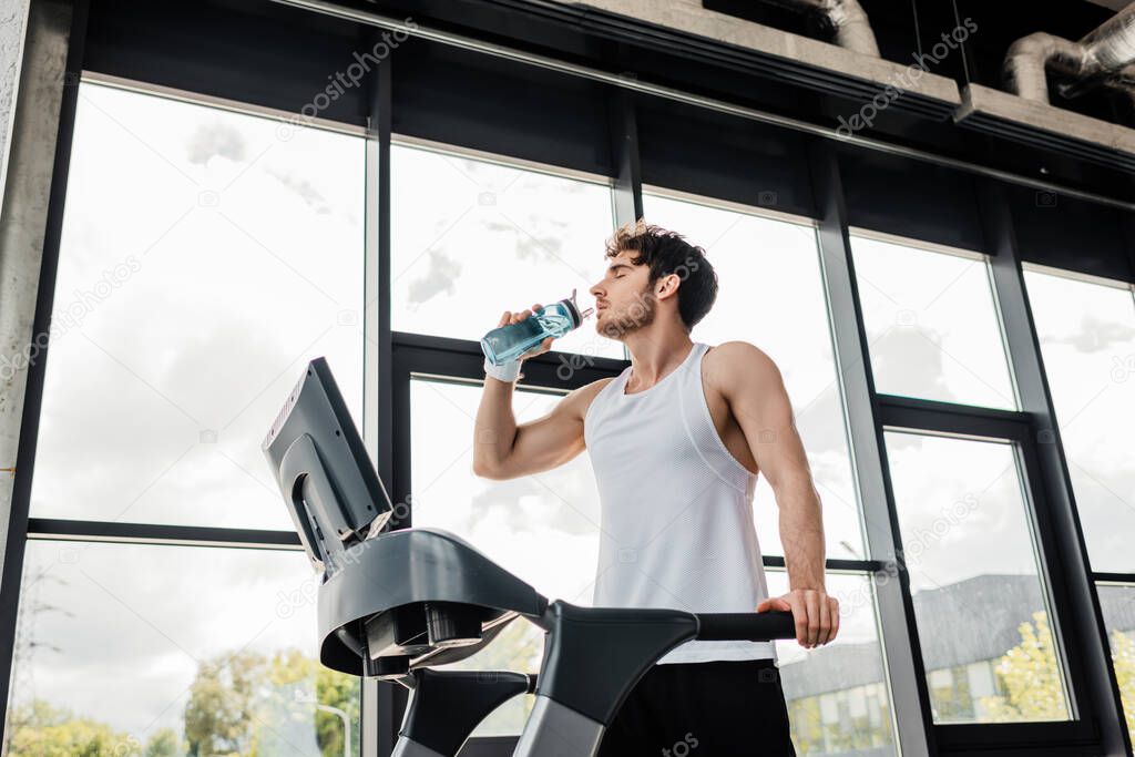low angle view of tired sportsman drinking water while standing on treadmill 