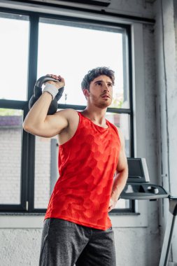 Sportif bir adam, elleri kalçasında dururken dambılla çalışıyor. 