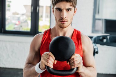 Atletik adam kameraya bakıyor ve spor merkezinde ağır bir halterle egzersiz yapıyor. 