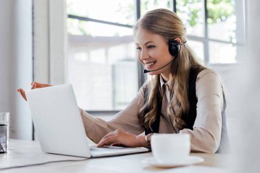 Laptop ve fincanın yanında gülümseyen kulaklıklı neşeli operatörün seçici odağı