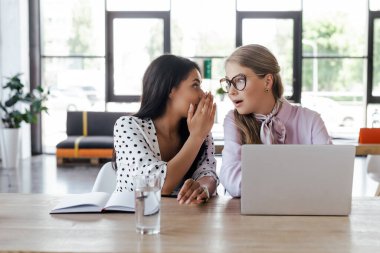 businesswomen gossiping near laptop in modern office clipart