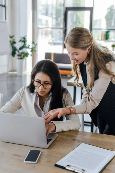 Gözlüklü çekici iş arkadaşının yanında duran ve dizüstü bilgisayarını işaret eden güzel bir iş kadınının seçici odağı 