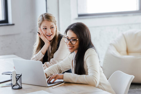 selective focus of excited businesswoman looking at happy coworker using laptop in office 