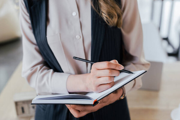 cropped view of businesswoman writing in notebook while standing in office