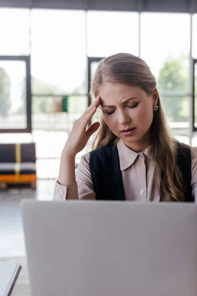 selective focus of tired businesswoman touching head near laptop