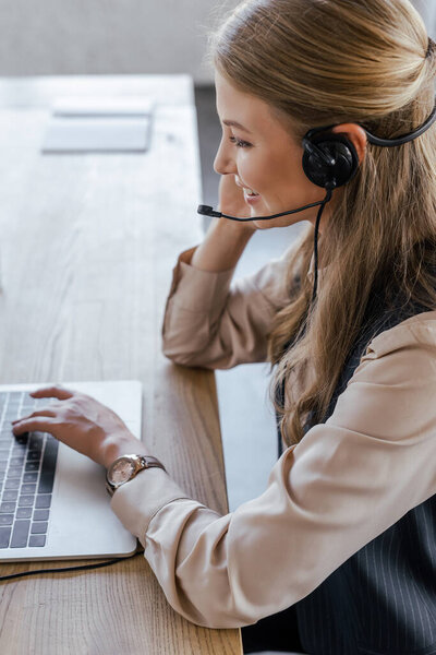 вид сбоку веселого оператора в наушнике, улыбающегося возле ноутбука
 