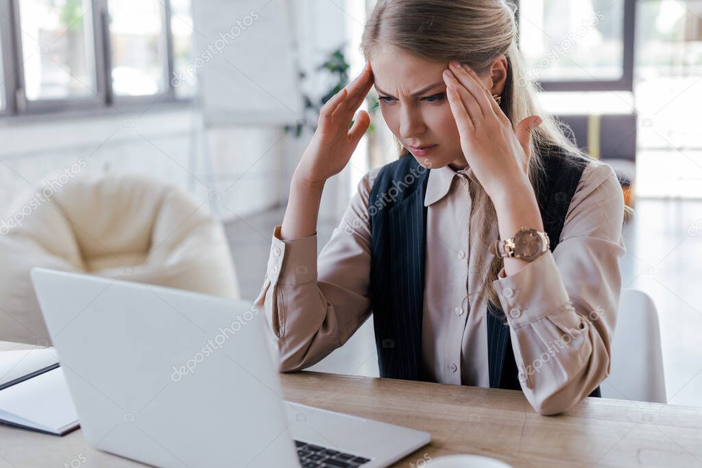 selective focus of exhausted businesswoman touching head near laptop 
