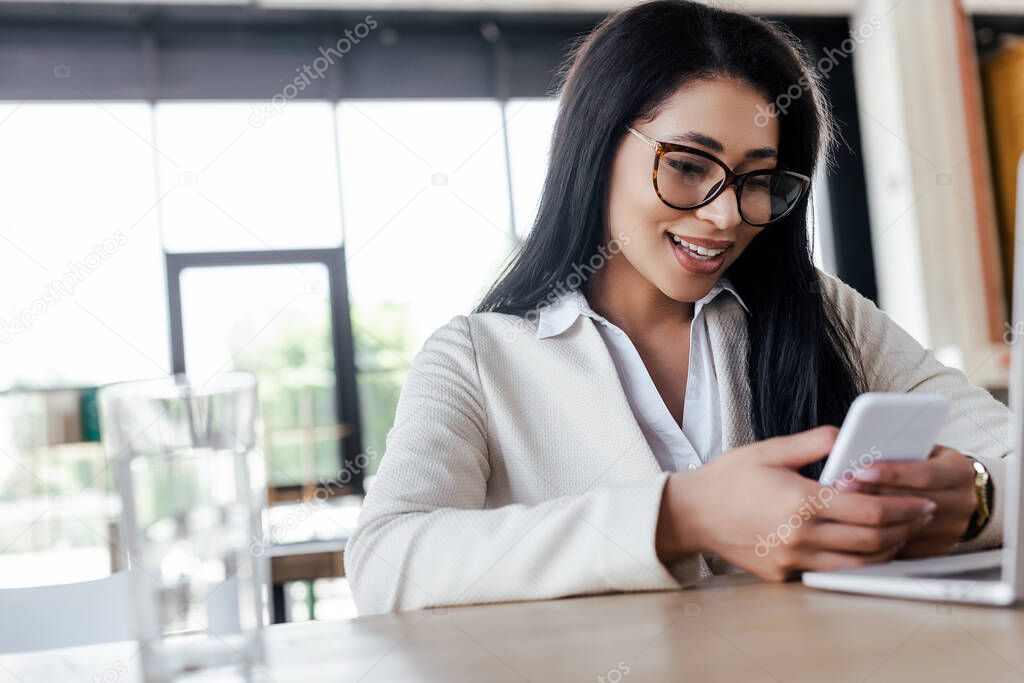 selective focus of happy businesswoman using smartphone near laptop 
