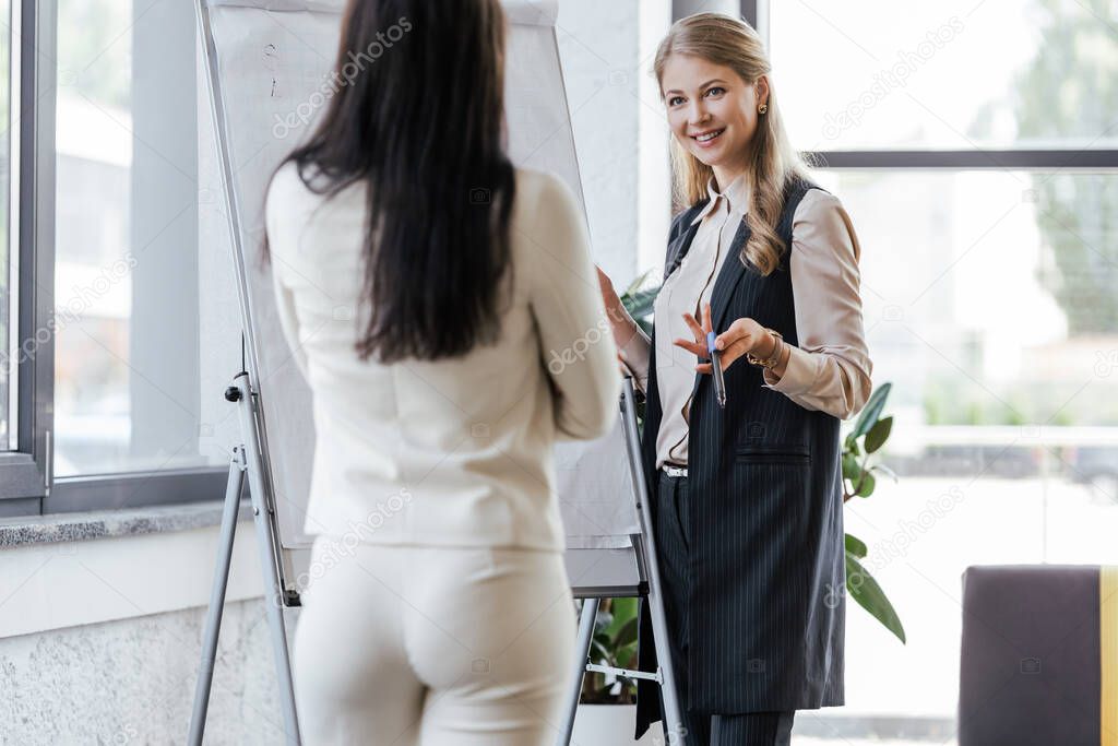 selective focus of happy businesswoman holding marker pen near flipchart and looking at coworker 