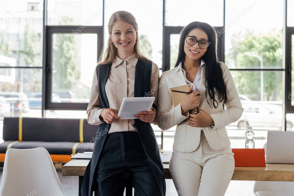 happy businesswoman holding digital tablet near beautiful coworker in glasses with notebook and pen