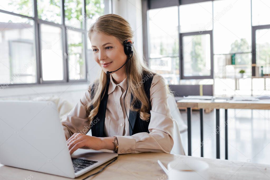 selective focus of happy operator in headset looking at laptop near cup