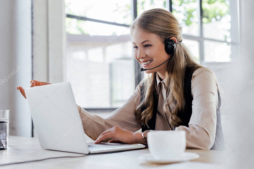 selective focus of cheerful operator in headset smiling near laptop and cup