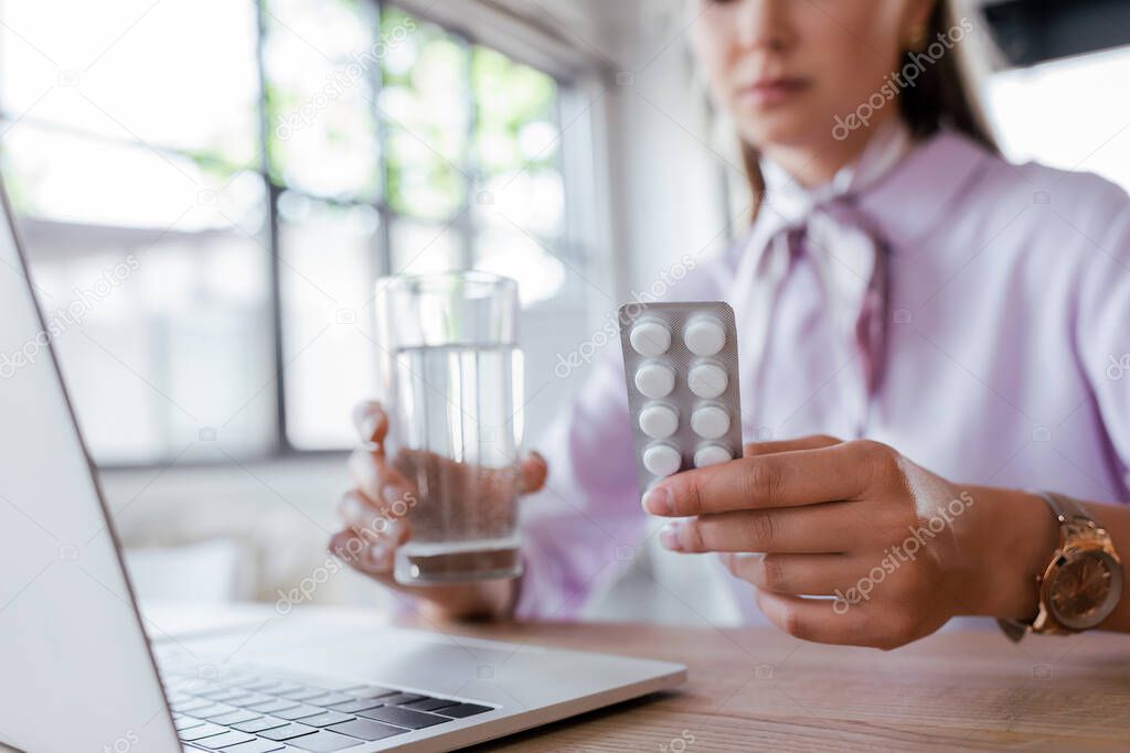 cropped view of woman holding glass of water and blister pack with pills near laptop 
