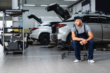 Atölyedeki arabaların yanında oturan ve kağıt bardak tutan bir tamirci. 