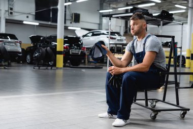 tired technician in overalls holding cap and gloves while sitting near cars in workshop  clipart