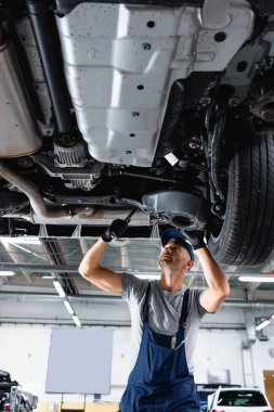 Servis istasyonunda araba tamir ederken İngiliz anahtarı tutan kapaklı ve tulumlu teknisyenin düşük açılı görüntüsü 