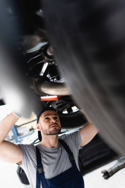selective focus of mechanic in cap holding flashlight while repairing car in service station  clipart