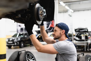 side view of mechanic in rubber gloves touching car brake and fixing vehicle  clipart