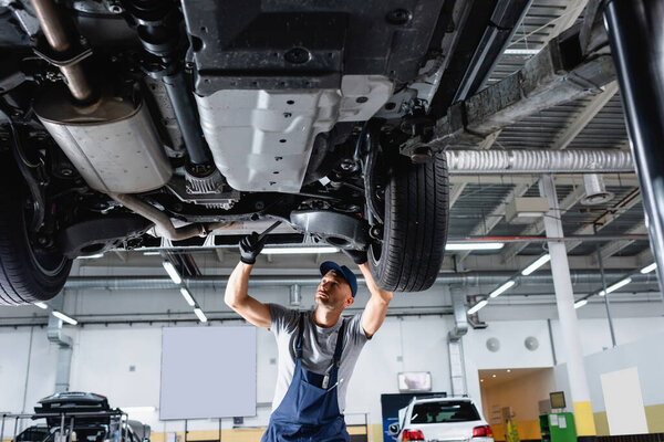low angle view of mechanic in cap and overalls holding wrench while repairing car in service station 