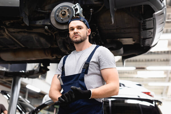 low angle view of mechanic in rubber gloves looking at camera while standing near car