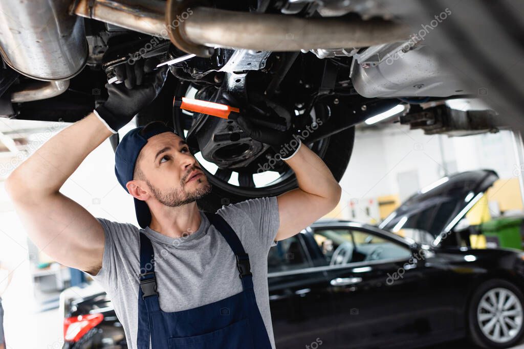 handsome mechanic in cap holding flashlight and wrench while repairing car in service station 