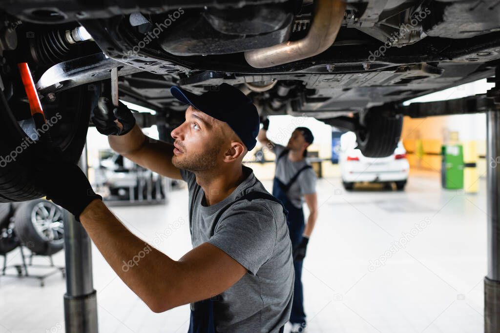 selective focus of handsome mechanic in cap holding flashlight and wrench while fixing car near coworker in service station 