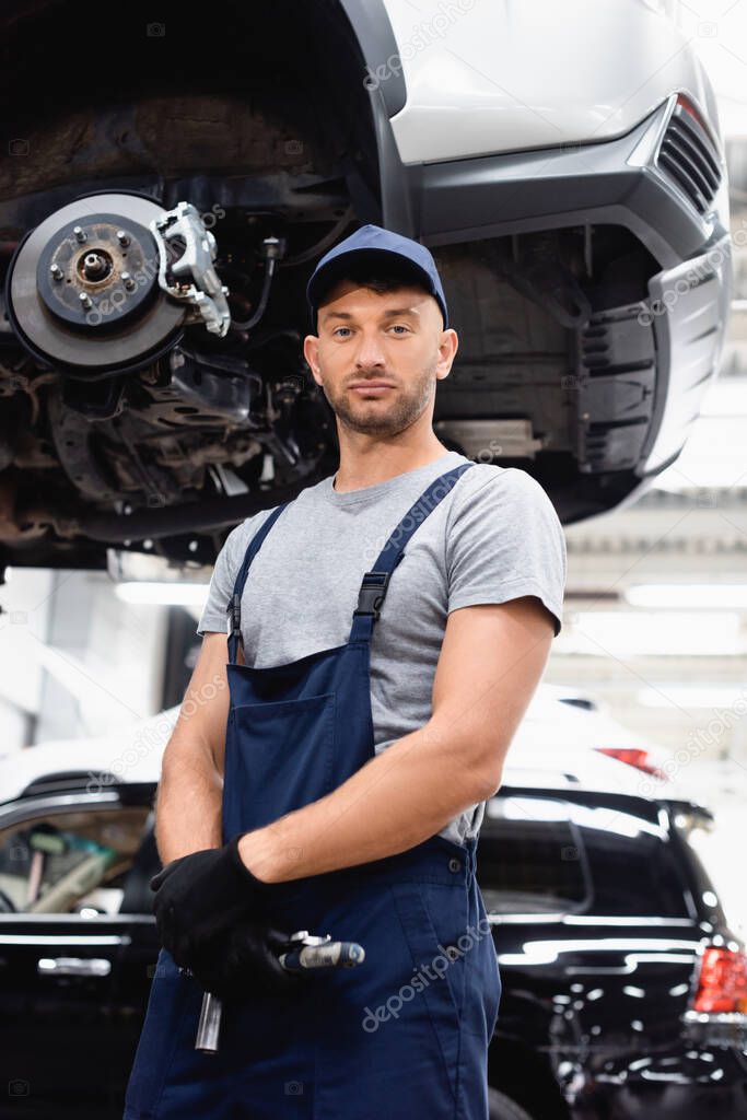 low angle view of mechanic in rubber gloves and cap looking at camera while standing near car