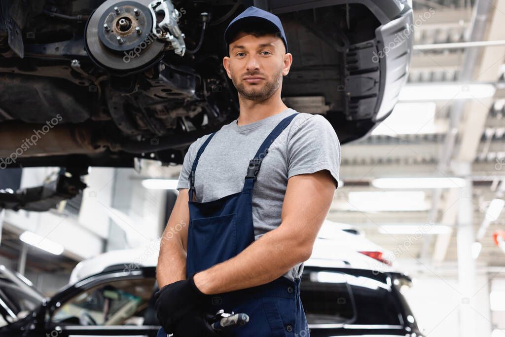 low angle view of mechanic in rubber gloves and overalls looking at camera while standing near car