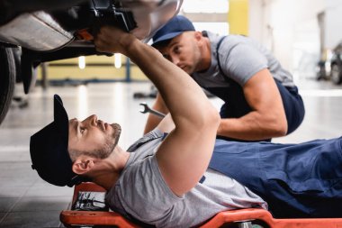 selective focus of mechanic in uniform lying and repairing auto near coworker in car service  clipart
