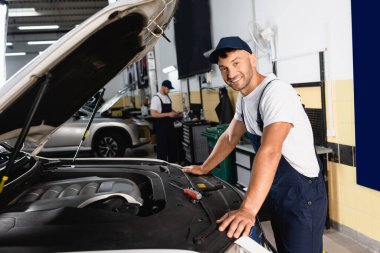 selective focus of happy mechanic in cap smiling near car and coworker in workshop  clipart