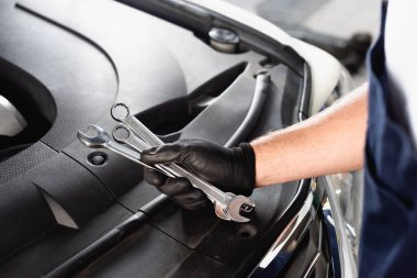 Cropped view of auto mechanic in latex glove holding wrenches near open hood clipart