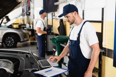 Servis istasyonundaki panonun yanındaki arabada dizüstü bilgisayar kullanan otomatik tamircinin seçici odak noktası