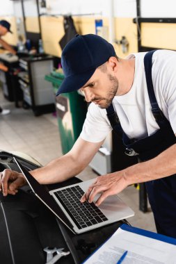 Auto mechanic typing on laptop on car near clipboard and wrenches at service station clipart