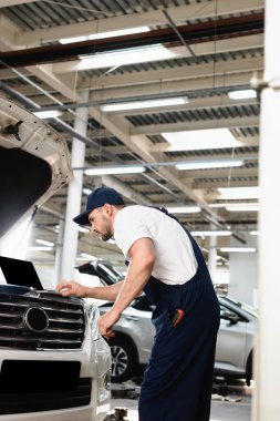 Auto mechanic leaning forward and typing on laptop on open hood of car at service station clipart
