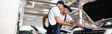Panoramic shot of auto mechanic holding laptop near open hood of car with colleague at service station clipart