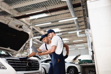 Side view of auto mechanic holding laptop near open hood of car with colleague at service station clipart