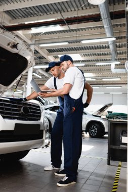 Servis istasyonunda iş arkadaşıyla birlikte açık kaputun yanında dizüstü bilgisayar tutuyor.