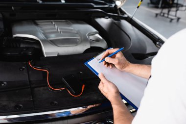 Cropped view of auto mechanic writing on clipboard near open hood of car with multimeter at service station clipart