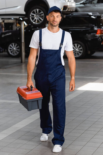 handsome mechanic in uniform and cap holding toolbox in service station