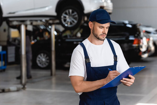 handsome mechanic in overalls and cap writing while holding clipboard